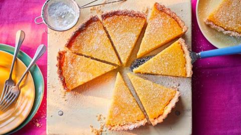 A lemon tart cit into slices and placed on a board with plates and forks alonside