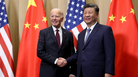 U.S. President Joe Biden and Chinese President Xi Jinping shaking hands in front of a backdrop displaying the flags of the United States and China. Biden is dressed in a dark suit with a white shirt and a burgundy tie, while Xi wears a navy suit paired with a white shirt and a blue tie.