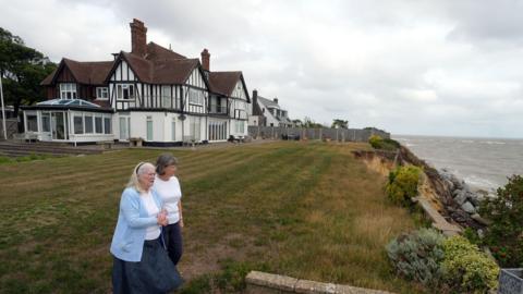 Jean Flick stands in her garden with her daughter at the edge of a cliff that overlooks the sea on a cloudy day. 