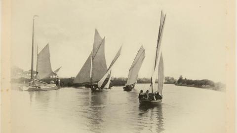 Sepia photo of boats being sailed on the Norfolk broads. There are four vessels each with a sail. You can see there are three people in the boat closest to the camera. 