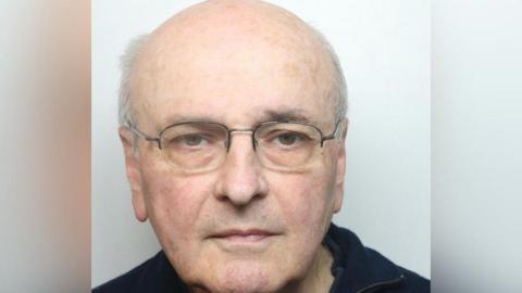 A mugshot of a man with white hair, silver-framed glasses and a black top.
