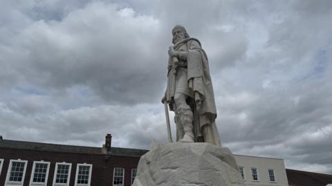 The statue of King Alfred is pictured on a plinth and taken from the ground nearby. A roof is visible in the background the skies are grey and overcast. 