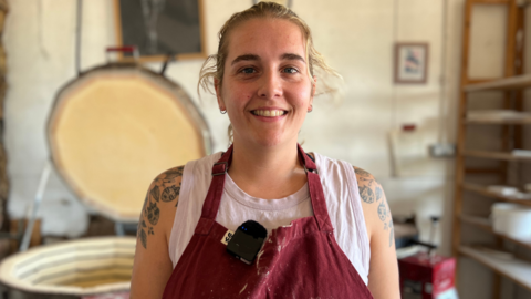 A woman wearing burgundy dungarees in a pottery studio.