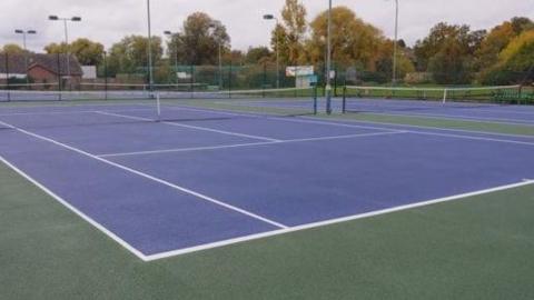A blue tennis court with nets set up. The surround of the court is green.