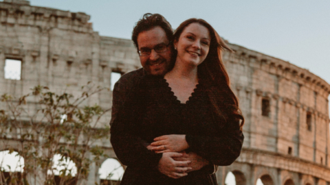 Megan McCley facing the camera while Dimitar Kashchive, standing behind her, rests his head on her shoulder. Their hands are touching around her waist and they are both smiling and wearing dark clothing. Behind them is the ruins of what looks like a coliseum.