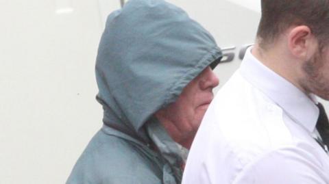 Alan MacPherson is wearing a blue waterproof jacket with the hood up as he is led away by a custody officer 