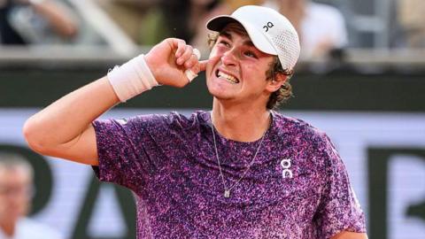 Joao Fonseca, wearing a purple top with white flecks and a white cap, grins and puts his right hand behind his ear to appreciate the support of the crowd