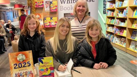 Tasha Ghouri sits at a table signing a copy of her book, with young fans Cassie and Reeva stood at either side.