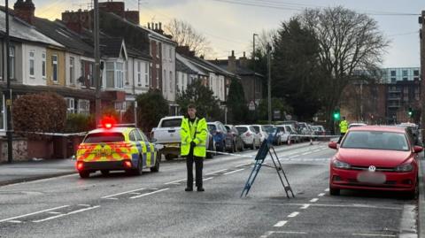 Two police officers wearing hi-vis uniforms stand at a road cordon on Carholme Road. There is a police car with its lights on and a road closure sign.