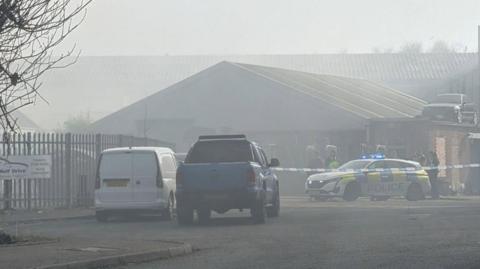 An industrial estate lies beyond police tape and a police car, with smoke filling the air and making for a hazy scene, partially obscuring other vehicles