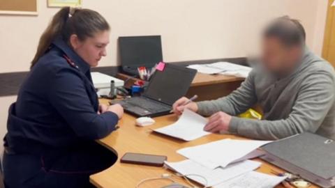 A police officer in blue uniform sits opposite a man in a grey sweater and blurred face at a table. The man is reading a sheet of paper, with a pen held in his right hand. Other pieces of paper are scattered on the table, where a mobile phone and charging lead, two laptops and thick lever-arch file can be seen. 