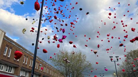 Lots of balloons, which are mostly red or pink, rise up to a cloudy sky having been let off by people below.