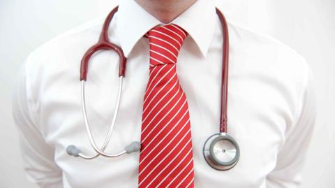 The chest of a doctor wearing a white shirt with a red striped tie and a stethoscope around his neck.