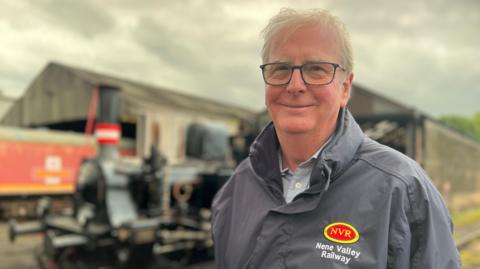 Michael Purcell standing in front of a locomotive and train sheds. He is wearing a grey raincoat with the railway's red and yellow branding on the left. He is smiling at the camera. He has square-framed glasses and short white hair.