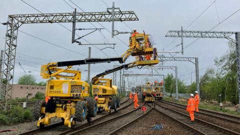 People in orange high vis jackets and trousers stand along a railways line. Four yellow cherry pickers are raised up to the railways power lines, people in the baskets are working on the lines.