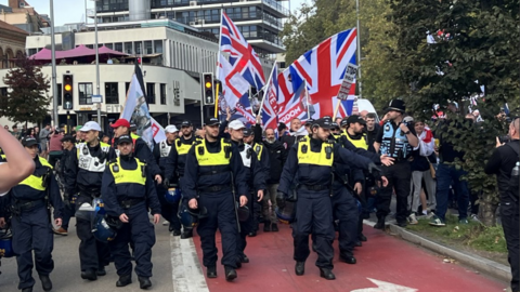 A line of police in police baseball caps and with riot gear attached to their belts march in front of a group of anti-immigration protesters waving Union Flags and St George's crosses