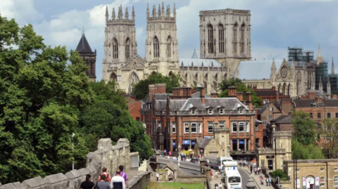 This is an image of York city centre, and is dominated by The Minster. The city walls, with people walking along them, are in the left of the shot. Lendal Bridge, to the right of the picture, is busy with traffic and people. The Minster's towers are framed by clouds and blue sky.