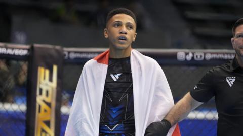 Cairo Thomas pictured in an MMA fighting ring either before or after a competition. He has a white towel wrapped around his shoulders. A coach or a referee stands next to home whilst holding his arm.