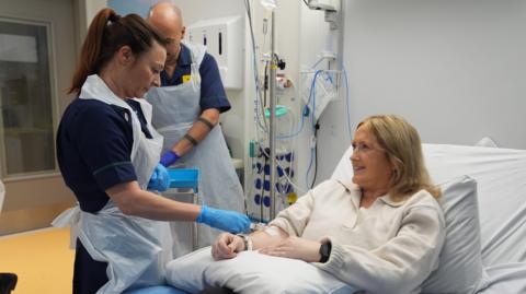 Katie Tinkler in a hospital bed with two medics standing alongside and one injecting something in to the patient's arm. Katie is looking up at the medic, who is wearing a dark uniform with plastic apron over the top.