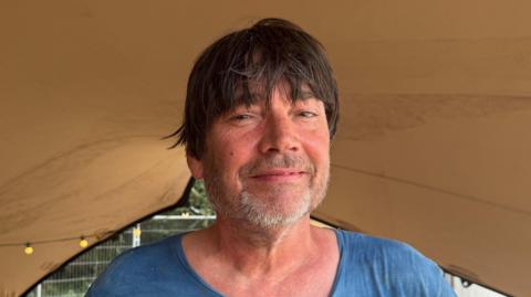 A head and shoulders image of Blur's Alex James. He is wearing a blue T-shirt and smiling while looking into the camera.
