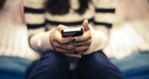 A girl wearing a black and white top, holds a mobile phone as she sits on a bed