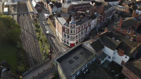 An aerial drone shot of a large red brick corner building with white frontage and windows. It has a clock tower and a spire. To its left are railway tracks