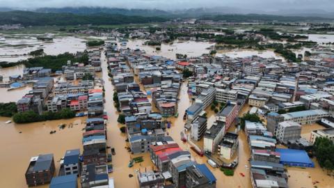 Aerial view of homes and buildings submerged in brown flood waters in Hengzhou, Nanning, China with a green mountain range and clody skies in the background
