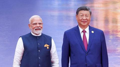 Narendra Modi and Xi Jinping smile for the cameras on stage against a shimmering blue and orange backdrop. Modi is wearing a blue vest over a white kurta with a golden pocket handkerchief, Xi is wearing a navy blue suit and a maroon tie.