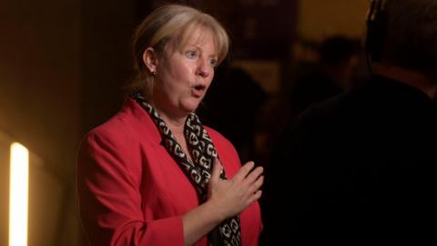 Shona Robison, wearing a red jacket, spkeaing to the media in the Scottish Parliament after delivering her Budget speech