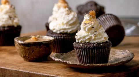 A chocolate cupcake on a rustic-looking ceramic plate, sat on a wooden table. It has white icing piled up on top of it in a swirl and bits of caramel and chocolate sprinkled over it. In the background are a few similar cupcakes which are blurred.