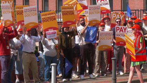A group of people holding up banners as doctors strike outside a hospital. Several people are wearing orange hats and hi-vis orange jackets. Some of the signs read "£18.62 is not a fair wage for a resident doctor".