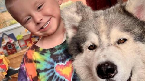 Three-year-old Ely stood next to Thunder. He has very short hair and is wearing a multicoloured T-shirt with a red and yellow heart in the middle. He has a huge grin and is looking directly at the camera. They are at Ely's home with toys in the background.