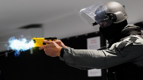 A side-on view of a prison officer firing a yellow Taser. The officer is wearing all-dark clothes, a black balaclava and a helmet with a shield screen which is up