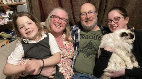 (L-R) Sophie, Mhari, Bill and Jess at home. They are sitting on a sofa and smiling at the camera. There is a curtain behind them.