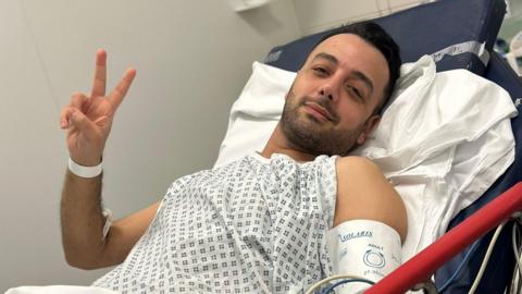 Pouria Zeraati lies in a hospital bed, wearing a hospital gown and wrist band, looking at the camera with two fingers raised in a peace sign