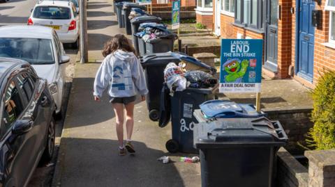 A woman walks down a Birmingham street seen with overflowing wheelie bins on one side of the pavement and parked cars on the other