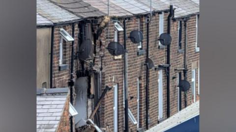 The image shows the rear side of a row of older brick terraced houses, viewed from an elevated or slightly angled perspective. The focus is on a section of severe structural damage affecting one of the properties.
In the centre of the image, one house has extensive vertical damage running from the roofline downward.
A large section of the brick wall has collapsed or been blown outward, exposing the interior structure.
