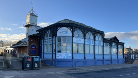 Blue fronted wooden cafe with intricate ironwork decorative framework. It has a Fish and Chips sign on the front. There are 2 bins standing to the left of the cafe which is near a road and there are steps either side. Behind the cafe building is an ornate Victorian clock tower.