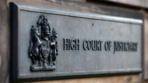 A sign attached to a wall which says High Court of Justiciary with a crest