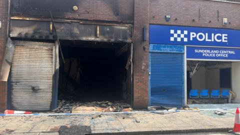 The burnt-out Citizens Advice branch. It has been blackened by flames and smoke. The window and front shutters have been destroyed. Next to it is a police office.