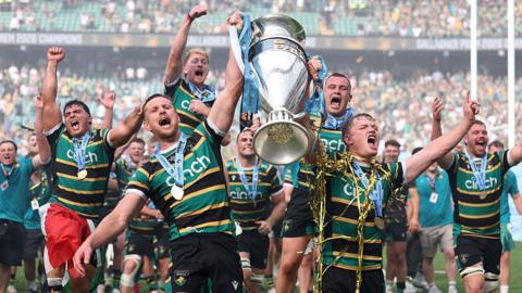 Northampton Saints players cheer as they carry a huge trophy, they are wearing medals and gold confetti