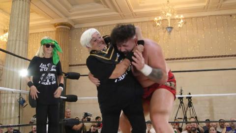 Two women wearing black t-shirts are in a wrestling ring. One of the women is holding a male wrestler with red shorts in a headlock. In the background there are columns, chandeliers and fairy lights covering the cream walls. There are also cameras and members of the audience watching on.  