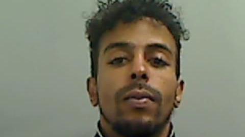 Mugshot of Amin Samater who has curly, black hair, a moustache and slight beard and very long eyelashes. 