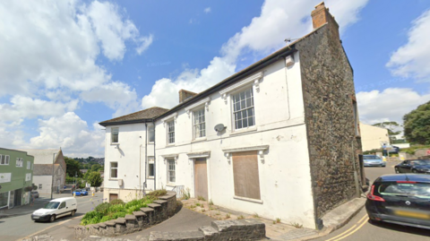 A street-view of a white derelict building. The windows are boarded up with wooden panels. It is on the corner of a street and roads either side.