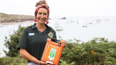Grace stood holding the orange defibrillator. She is stood in front of a sea view which has boats on the water. 