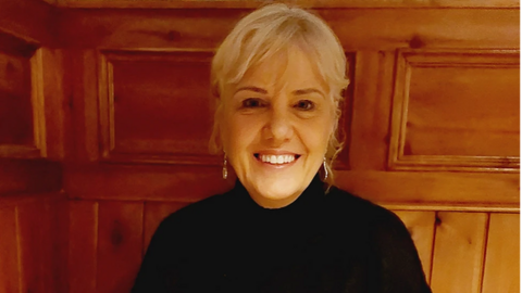 Jenny Dunlop. She has blonde hair tied back with a fringe, wearing earring and a black top. Wood panelling is behind her.