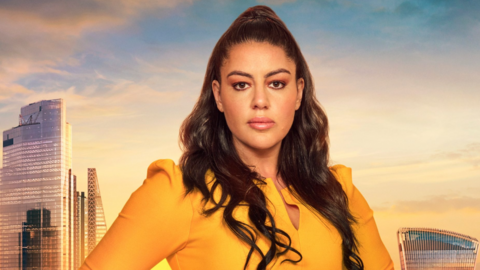 Picture of Nadia Suliaman in a mustard yellow top. She stands infront of a green screen of London. She looks blankly into the camera and has brown eyes and dark brown hair. 