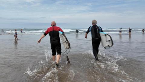 Two men in wetsuits are walking into the sea at Bude, Cornwall. they are both holding surf boards and