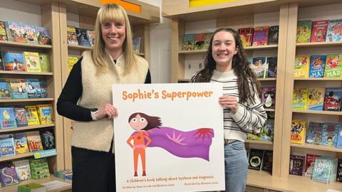 Dawn Crowle and Rhiannon Jones. Dawn has long blonde hair and is wearing black with a cream knitted vest. Rhiannon has long dark hair and is wearing blue jeans and a striped jumper. They are stood side by side, holding a large copy of the Sophie's Superpower book cover. It is white with an illustrated superhero wearing an orange suit with a purple cape. 