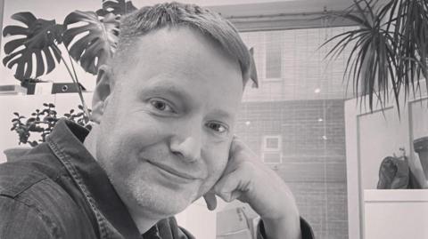 Chris with his head resting on his hand smiling at the camera with a plant in the background